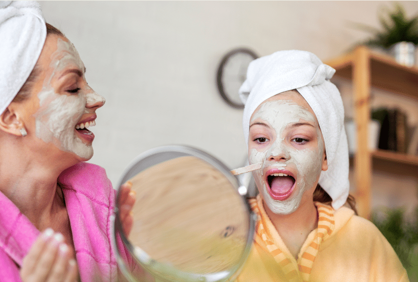 Two people with white facial masks and towels wrapped around their heads smile and laugh together. One holds a mirror, while the other has a clothespin on her nose. Both wear colorful bathrobes.