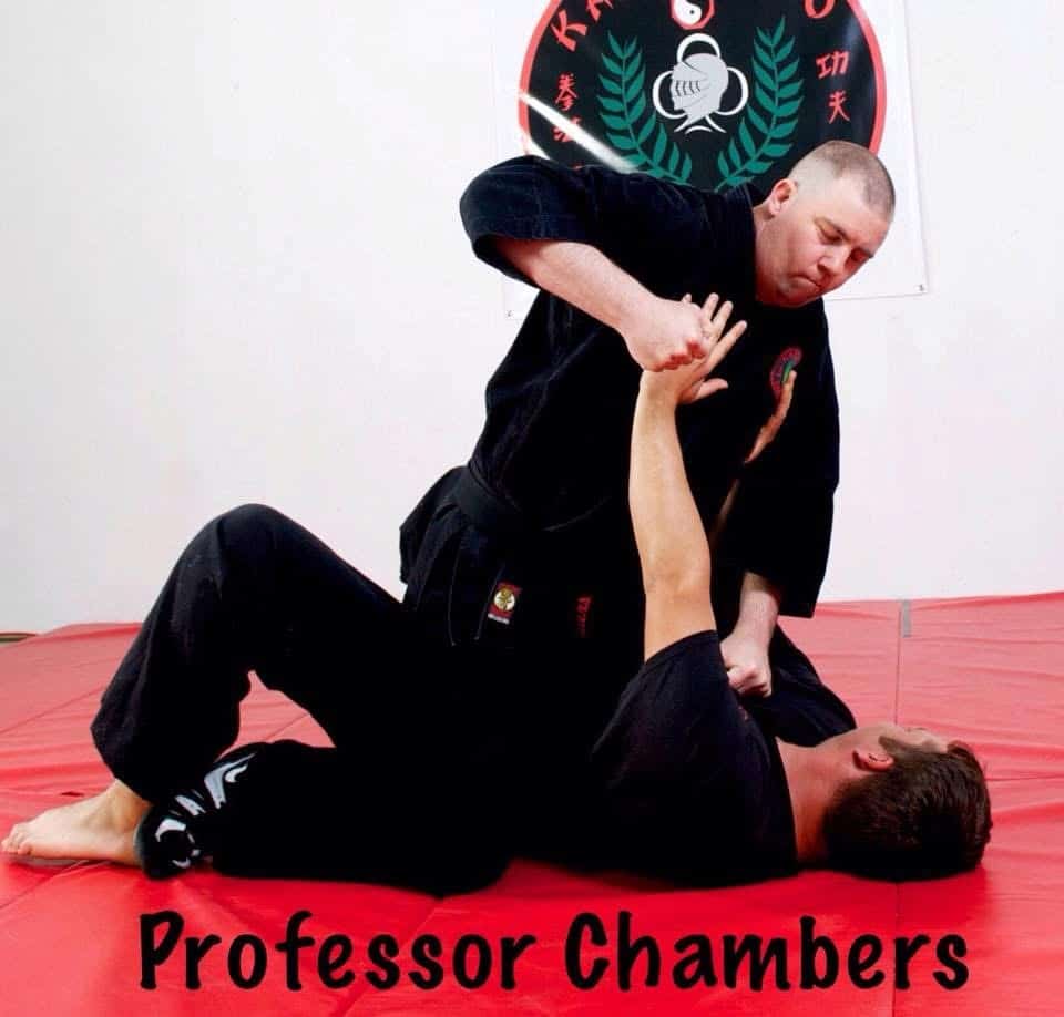Two people practice martial arts on red mats. One person is pinning the other to the ground, preparing to strike. Both wear black uniforms. The words Professor Chambers are written at the bottom of the image.